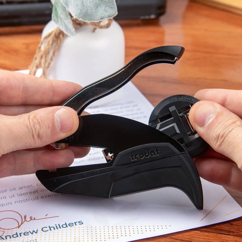 Hands demonstrating how to insert or remove the interchangeable embosser plate on a black Trodat embossing tool